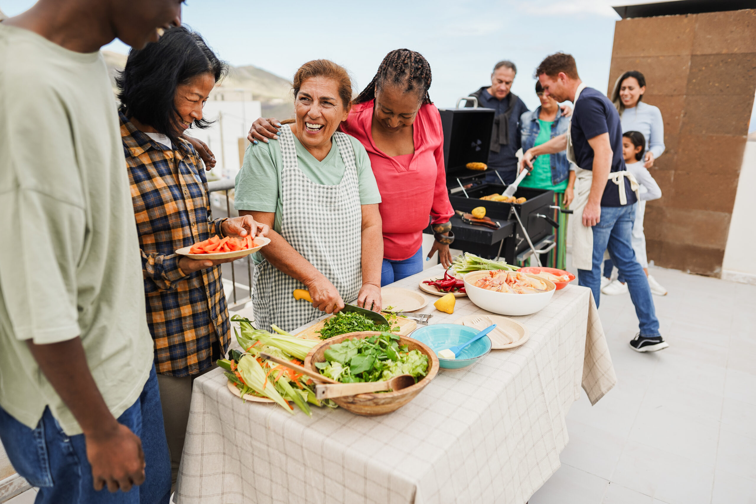 Multi generational people doing barbecue at home's rooftop - Multiracial friends having fun eating and cooking together during weekend day - Summer and food concept - Main focus on latin woman face